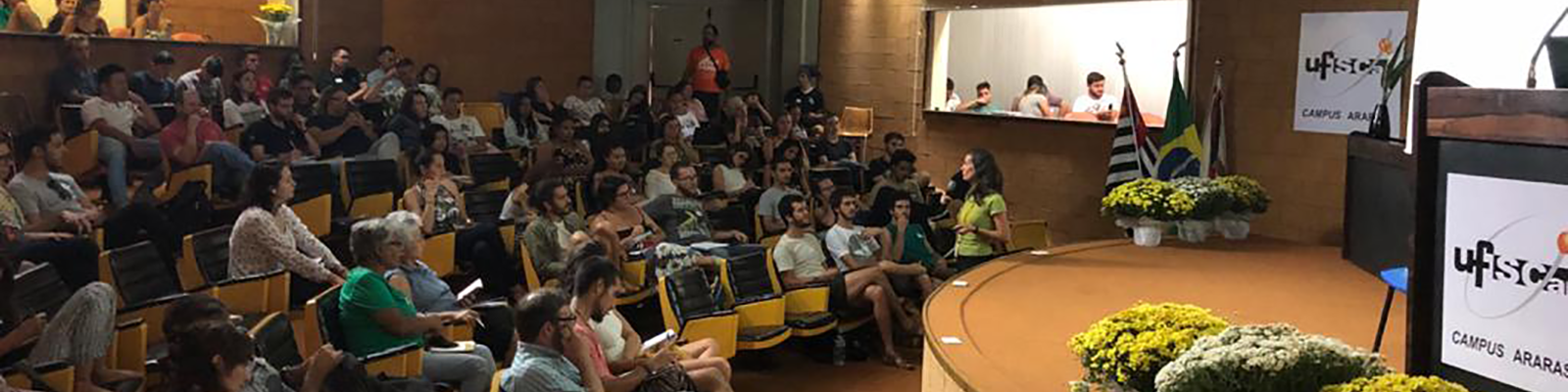 CCA-UFSCar’s amphitheater with people sitting on orange coloured chairs with black seats, fixed on the side, stage with pulpit and flower pots. On the stage’s edge, Brasil’s, São Paulo’s and Araras’ (municipality) flags. On the ceiling, spotlights turned to the stage.