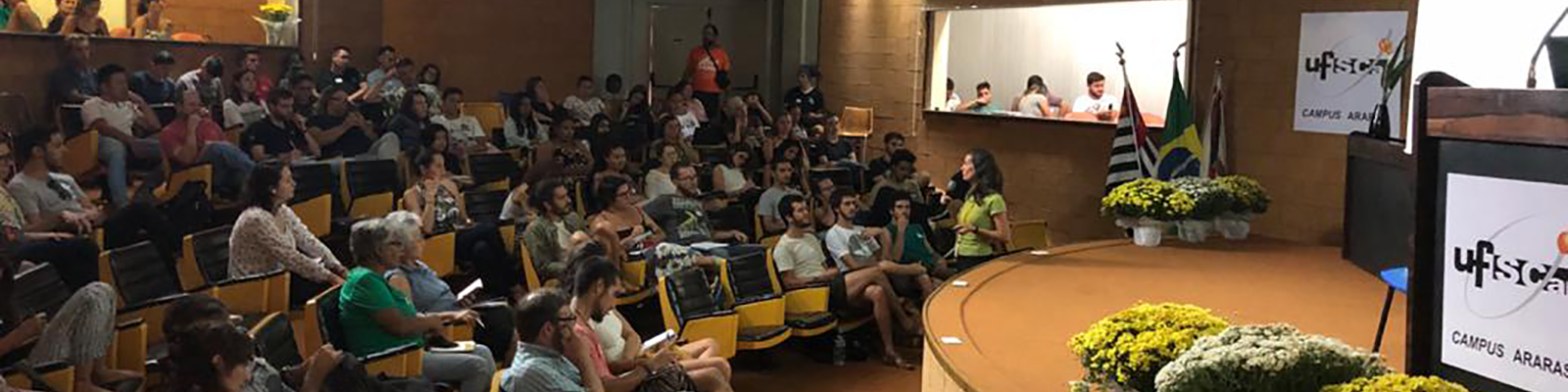 Anfiteatro del CCA-UFSCar con personas sentadas sobre las sillas, lateral en color naranja y asientos de color negro, escenario con púlpito y macetas, al final del escenario banderas de Brasil, estado de São Paulo y municipio de Araras. En el techo focos de luces dirigidos al escenario.