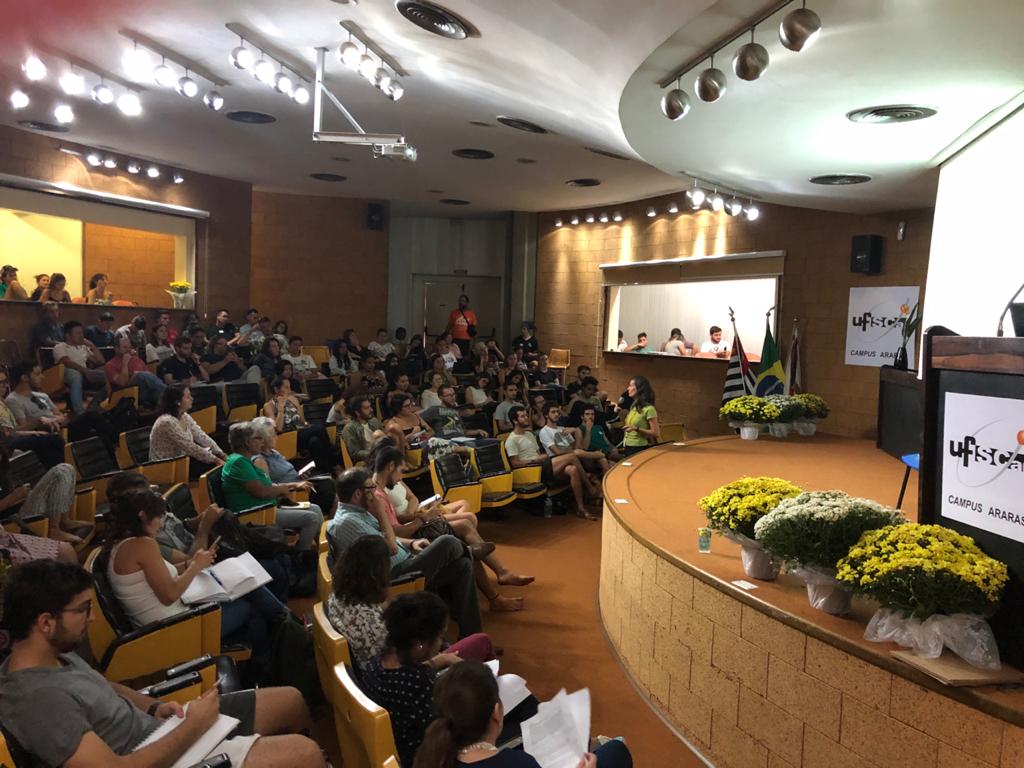 Anfiteatro do CCA-UFSCar com pessoas sentadas em cadeiras fixas lateral na cor laranja e o assento em preto, palco com púlpito e vasos de flores, no final do palco bandeiras do Brasil, estado de São Paulo e município de Araras. No teto holofotes de luzes dirigidos ao palco.