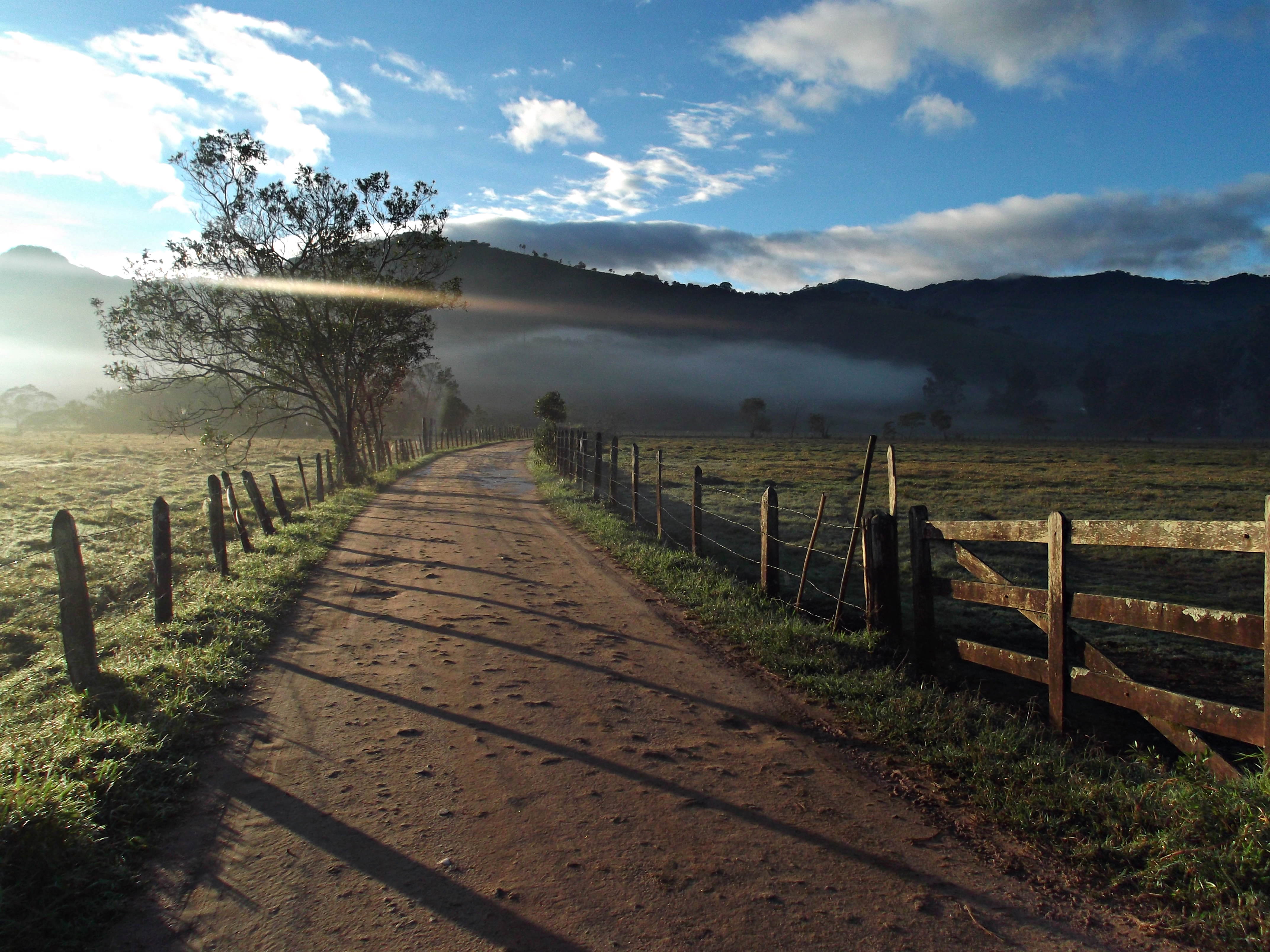 Foto do 1º Concurso de Fotografia- PPGADR - Seu Olhar Sobre o Ambiente. Caminho da roça. Ednilson M. de Lima e Silva. Estrada de terra batida ao lado cercas em madeira e arame, uma porteira do lado direito e algumas árvores do lado esquerdo, ao fundo névoa e um morro.