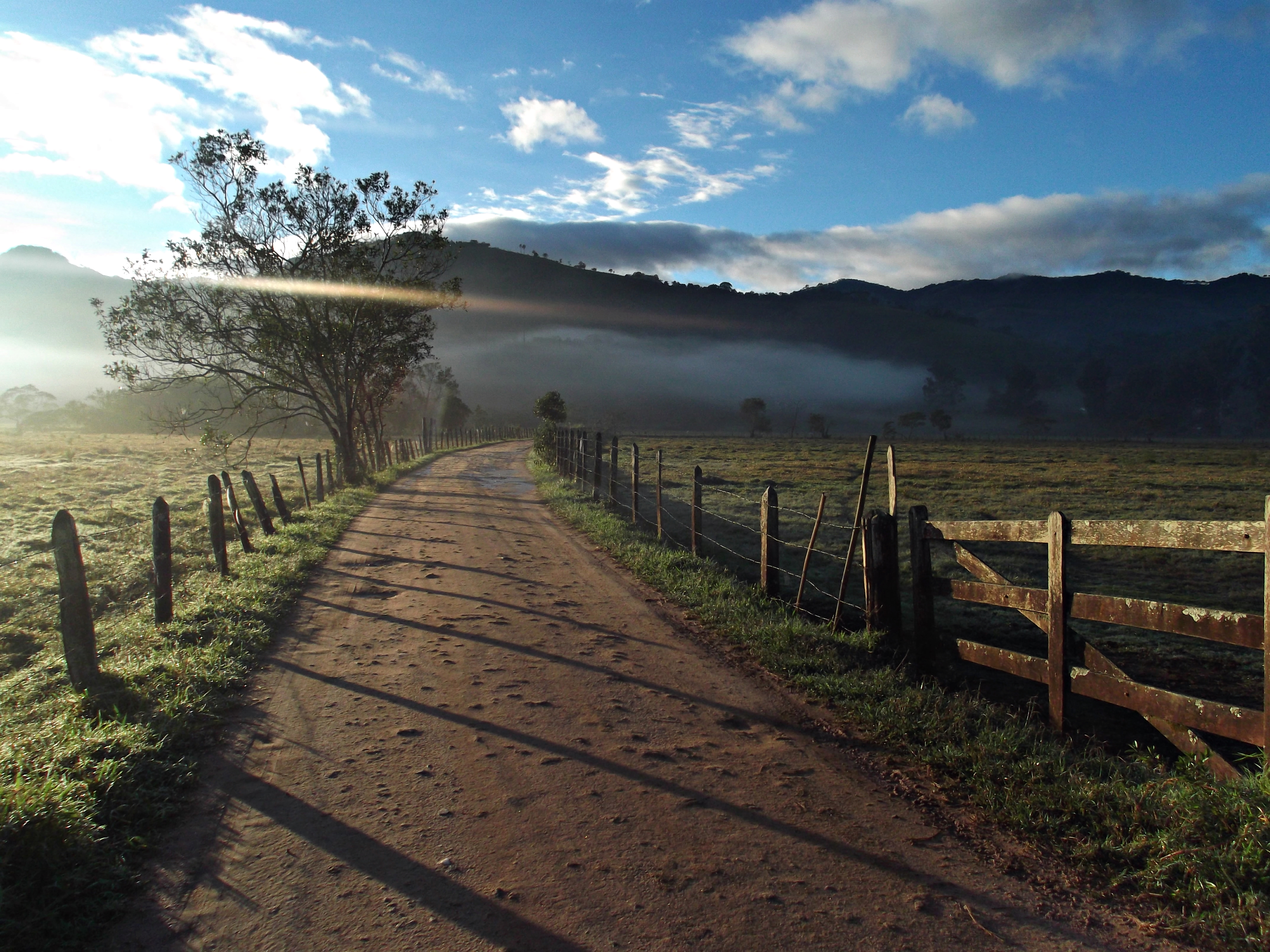 Foto do 1º Concurso de Fotografia- PPGADR - Seu Olhar Sobre o Ambiente. Caminho da roça. Ednilson M. de Lima e Silva.  Estrada de terra batida ao lado cercas em madeira e arame, uma porteira do lado direito e algumas árvores do lado esquerdo, ao fundo névoa e um morro.